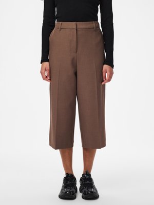 Osoba ubrana w brązowe szerokie, skrócone spodnie, czarną bluzkę z długim rękawem i czarne trampki, stojąca na tle białego tła.