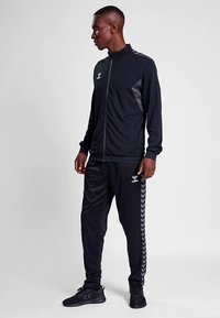 Sort atletisk træningssæt med en lynlåsjakke med opretstående krave og matchende tilspidsede bukser. Fremhævet med et Chevron-mønster på siderne.