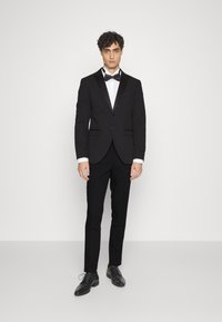 Tuxedo nero con rever a scialle, camicia bianca, papillon nero e pantaloni neri slim-fit. Indossato con scarpe eleganti nere. Sfondo semplice.
