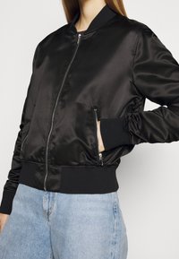 Veste bomber en satin noir avec fermeture éclair frontale et poches zippées portée sur un jean bleu clair, main dans la poche, vue recadrée.