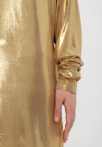 Metalni zlatni dolg rokav oblačilo z gladko teksturo. Manšeta je zbrana, kar ustvarja napihnjen učinek na zapestju.