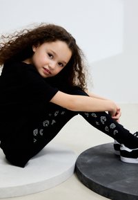 Črna majica s kratkimi rokavi in pajkice z vbodnim vzorcem, sedi na sivo-belih krožnih ploščadih. Obutev je črna z belimi podplati.