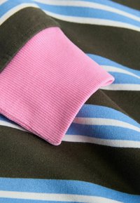 Pruhovaná látka s modrými, bílými a olivově zelenými pruhy. Žebrovaný růžový lem vytváří kontrast. Měkká texturovaná tkanina s uvolněným designem.