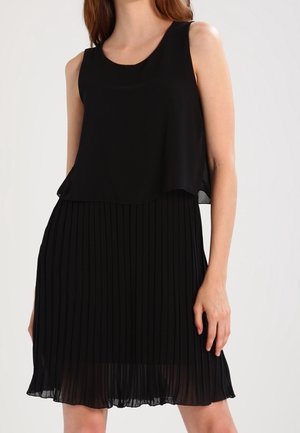 Ärmelloses schwarzes Kleid mit geschichtetem Chiffontop und plissiertem Rock, getragen von einer stehenden Frau vor einem einfarbigen Hintergrund.