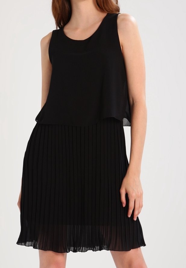 Robe noire sans manches avec haut en chiffon superposé et jupe plissée, portée par une femme debout sur un fond uni.