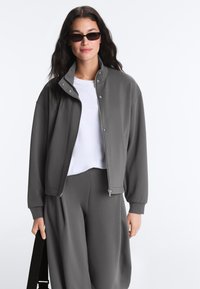 Femme aux longs cheveux foncés portant des lunettes de soleil noires, une veste grise zippée, un pantalon assorti et une chemise blanche, tenant une sangle de sac noire.