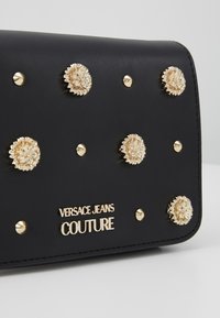 Czarna skórzana torebka ozdobiona złotymi ćwiekami w kształcie lwa oraz złotym napisem "Versace Jeans Couture".