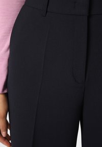 Marineblaue Hose mit glatter Textur und taillierter Passform, ausgestattet mit einer Fronttasche und einem leicht hohen Bund, kombiniert mit einem rosa Langarmoberteil.