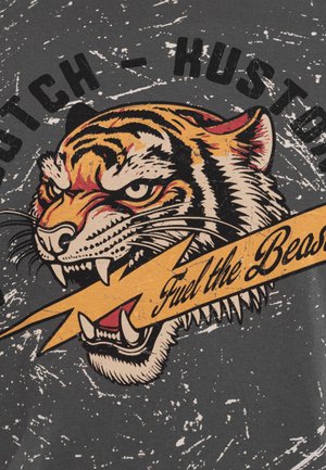 Tête de tigre aux dents découvertes mordant un éclair avec le texte "Fuel the Beast" sur un fond gris foncé usé.