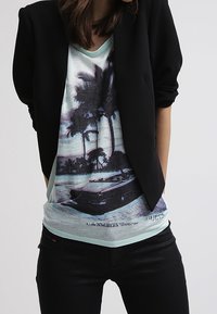 Sort blazer over en lysblå grafisk T-shirt med et palmetræ og bil-design. Sorte jeans fuldender outfittet og skaber et afslappet look.
