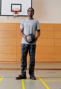 Basketballspiller i en grå Jordan t-skjorte, holder en svart Molten basketball, iført rutete svarte bukser og brune sko, står på en bane.