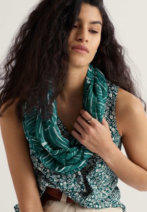 Sieviete ar gariem viļņainiem matiem valkā zaļu ziedotu bezroku topu un zaļu un baltu svītrainu šalli, tur šalli pie kakla.