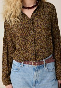 Chemise à imprimé léopard dans des tons marron et olive, dotée de manches longues et d'une coupe décontractée, associée à un jean bleu clair et une ceinture bordeaux.