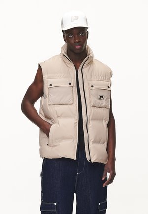 Beige puffer vest with zip front, two chest pockets, and a logo. Worn over a black shirt, paired with dark denim cargo pants.