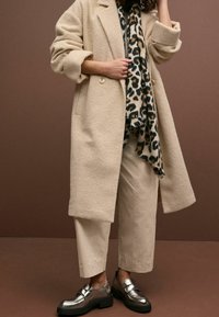 Manteau beige texturé avec manches retroussées, écharpe à motif léopard, pantalon crème et chaussures métalliques argentées sur un fond brun.