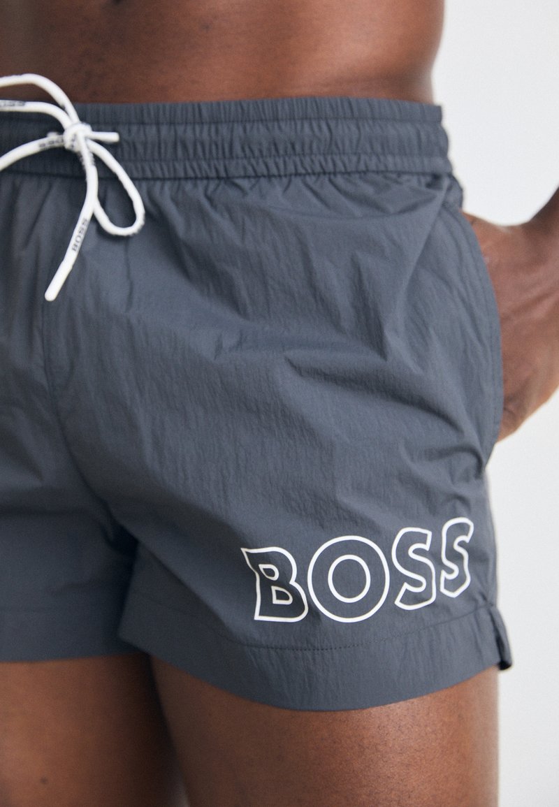 Szare kąpielówki wykonane z lekkiego materiału, z białym sznurkiem i słowem "BOSS" w wyrazistym nadruku po boku.