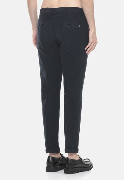 Uomo che indossa pantaloni in corduroy slim-fit blu navy scuro e mocassini in pelle nera, in piedi con le spalle rivolte verso la fotocamera su uno sfondo bianco.