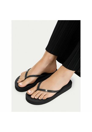Stopy w czarnych klapkach typu flip-flop z cienkimi paskami i srebrnymi akcentami, w połączeniu z czarnymi plisowanymi spodniami na jasnym tle.