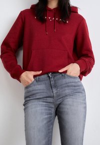 Rode hoodie met buideltas en trekkoord, met witte logotekst. Gecombineerd met lichtgrijze jeans met subtiele textuur en pasvorm.