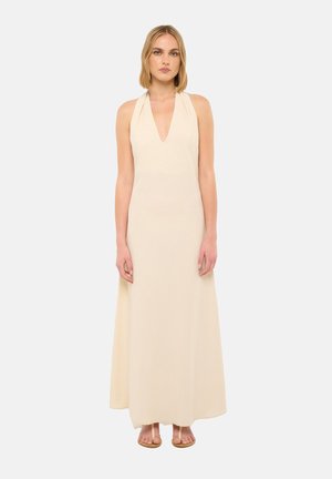 Donna con capelli biondi lisci, vestita con un abito crema senza maniche con scollo all'americana e sandali beige con punta aperta, in piedi davanti a uno sfondo semplice.