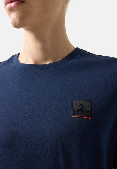 Gros plan d'une personne portant un t-shirt bleu marine avec un petit écusson logo noir et rouge "FIRE + ICE" sur la poitrine.