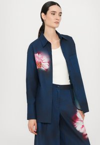 Camicia blu navy con bottoni, caratterizzata da una stampa floreale su un lato. Tessuto di cotone con una texture morbida, abbinata a pantaloni a gamba larga coordinati.