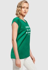 Grünes Baumwoll-T-Shirt mit entspannter Passform, kurzen Ärmeln und weitem Halsausschnitt. Mit einem weißen Textdesign auf der Vorderseite. Kombiniert mit weißen Hosen.