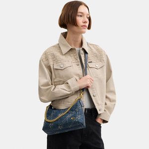 Mujer con chaqueta beige y pantalones negros, sosteniendo un bolso azul con cadena dorada sobre el hombro, mirando hacia un lado.