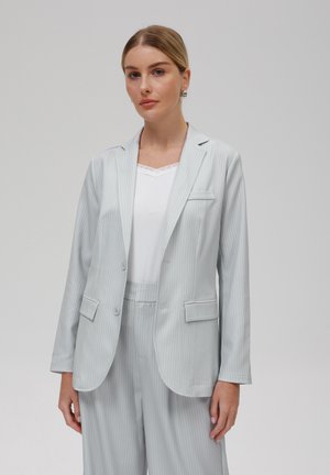 Blazer gris clair à rayures fines avec une coupe ajustée, deux boutons et poches avant. Porté par-dessus un top blanc avec dentelle. Tissu lisse et léger.