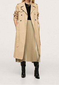 Manteau beige en trench avec des boutons double boutonnage, épaulettes, associé à une jupe plissée beige clair et des bottes noires montantes.