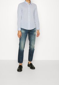 Man med en åtsittande blå och vit rutad långärmad skjorta, bleka blå jeans och svarta läderskor som står mot vit bakgrund.