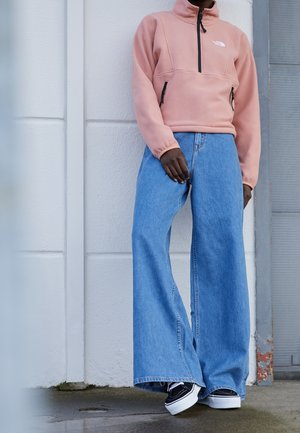 Personne portant une veste en polaire rose avec des poches avant, un jean bleu à jambes larges et des baskets noires et blanches, debout contre un mur blanc.