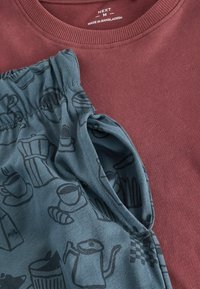 Tamsiai raudonas džemperis su austa apykakle ir trumpomis rankovėmis šalia žydrų kelnių, ant kurių yra juodai kontūruotas kavos motyvas.