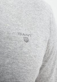Šedý pletený svetr s měkkou strukturou, na levé straně je umístěno malé vyšívané logo GANT v tmavě šedé barvě.