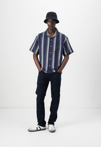 Cămașă cu mânecă scurtă, în dungi navy și crem, cu un buzunar; asortată cu pantaloni închiși la culoare și adidași albi cu accente negre.