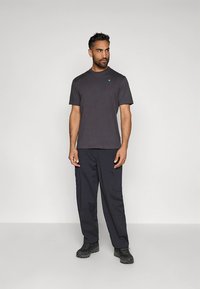 Donkergrijs heren T-shirt met korte mouwen en een turquoise logo op de borst, gecombineerd met donkerblauwe cargobroek en zwart schoeisel.