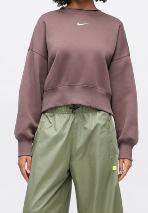 Person iført en lilla Nike sweatshirt og løse, højtaljede grønne bukser med snøre i taljen, står foran en ensfarvet baggrund.
