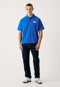 Blå pikétröja med krage, korta ärmar och "Red Bull Racing"-logotyp. Kombinerad med mörka jeans och vita sneakers. Enkla bakgrund.