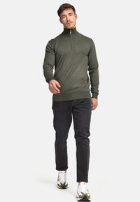 Olijfgroene ritssluiting sweatshirt met hoge kraag, slim-fit ontwerp, geribbelde manchetten en zoom, gecombineerd met donkere jeans en witte sneakers.