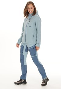 Ljusblå jacka med huva, framfickor med dragkedja och matchande blå byxor med flera fickor med dragkedja och färgblockering. Svarta vandringskängor.