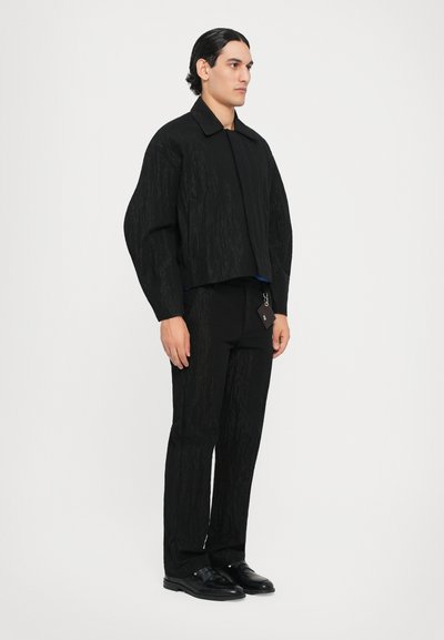 Schwarze, strukturierte Jacke mit einem taillierten Design, kombiniert mit passenden, schmal geschnittenen Hosen und schwarzen Schuhen. Verfügt über einen Kragen und minimalistische Hardware.