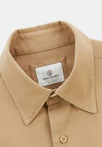 Chemise beige à manches longues avec un col classique, deux boutons au niveau du cou et une étiquette en tissu de marque visible à l'intérieur du col.