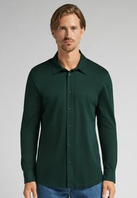 Hombre con cabello castaño claro que lleva una camisa de botones de manga larga verde oscuro y jeans azules frente a un fondo claro.