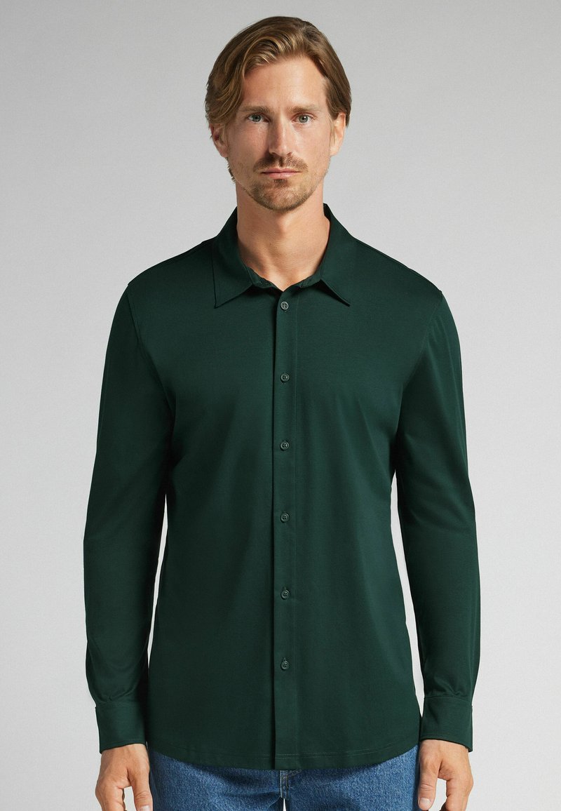 Hombre con cabello castaño claro que lleva una camisa de botones de manga larga verde oscuro y jeans azules frente a un fondo claro.