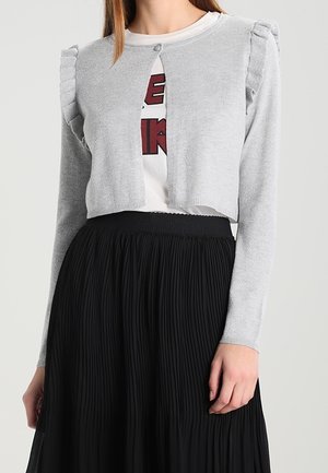 Cárdigan gris corto con hombros acampanados, cierre de botones, llevado sobre una camiseta blanca con gráfico, combinado con una falda negra plissada.