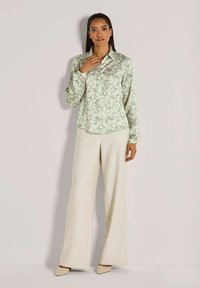 Vrouw die tegen een effen muur staat, gekleed in een lange mouwen paisley blouse, beige brede broek en beige puntige hakken.