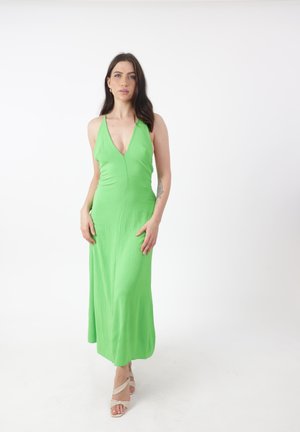 Donna con lunghi capelli scuri che indossa un vestito verde brillante senza maniche e sandali bianchi con cinturini, in piedi davanti a uno sfondo bianco.