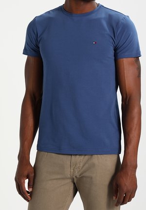 T-shirt bleu à col rond en coton doux, manches courtes, avec un petit logo rouge et blanc sur la poitrine gauche. Porté avec un pantalon beige.