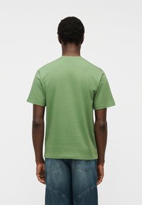 Άνδρας φορώντας μονόχρωμο ανοιχτό πράσινο κοντομάνικο μπλουζάκι και μπλε τζιν, στέκεται με την πλάτη προς τα εμπρός μπροστά σε φωτεινό φόντο.