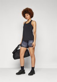 Top deportivo negro combinado con shorts azul y negro estampados, sosteniendo una bolsa de gimnasio negra. Los zapatos son negros con un diseño de malla texturizada.